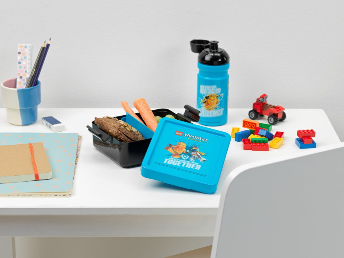 Niebieski lunchbox LEGO Ninjago z przekąskami i butelką na wodę na białym biurku do szkoły