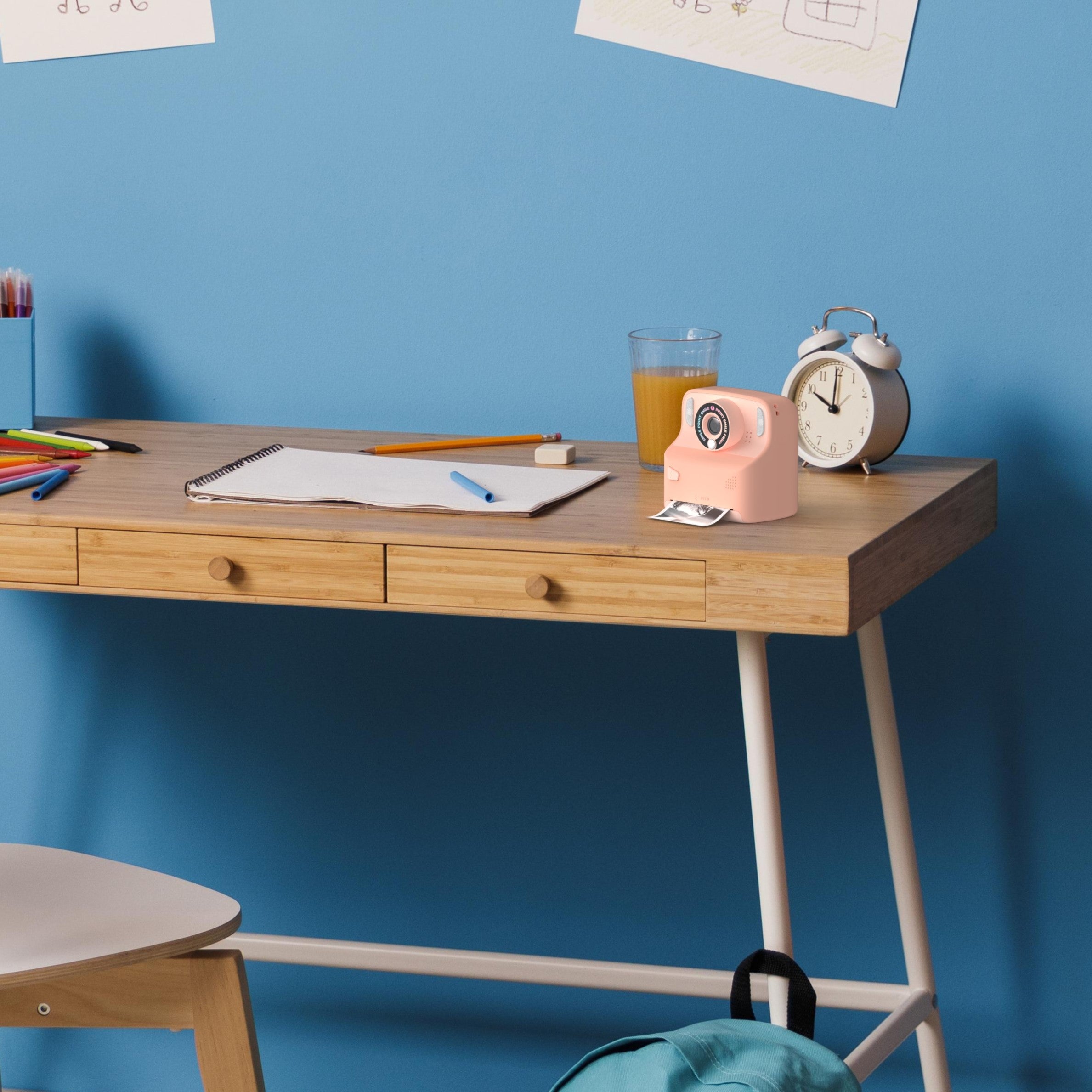 Różowy aparat cyfrowy z drukarką Yamann na biurku z notesem, zegarem i szklanką soku