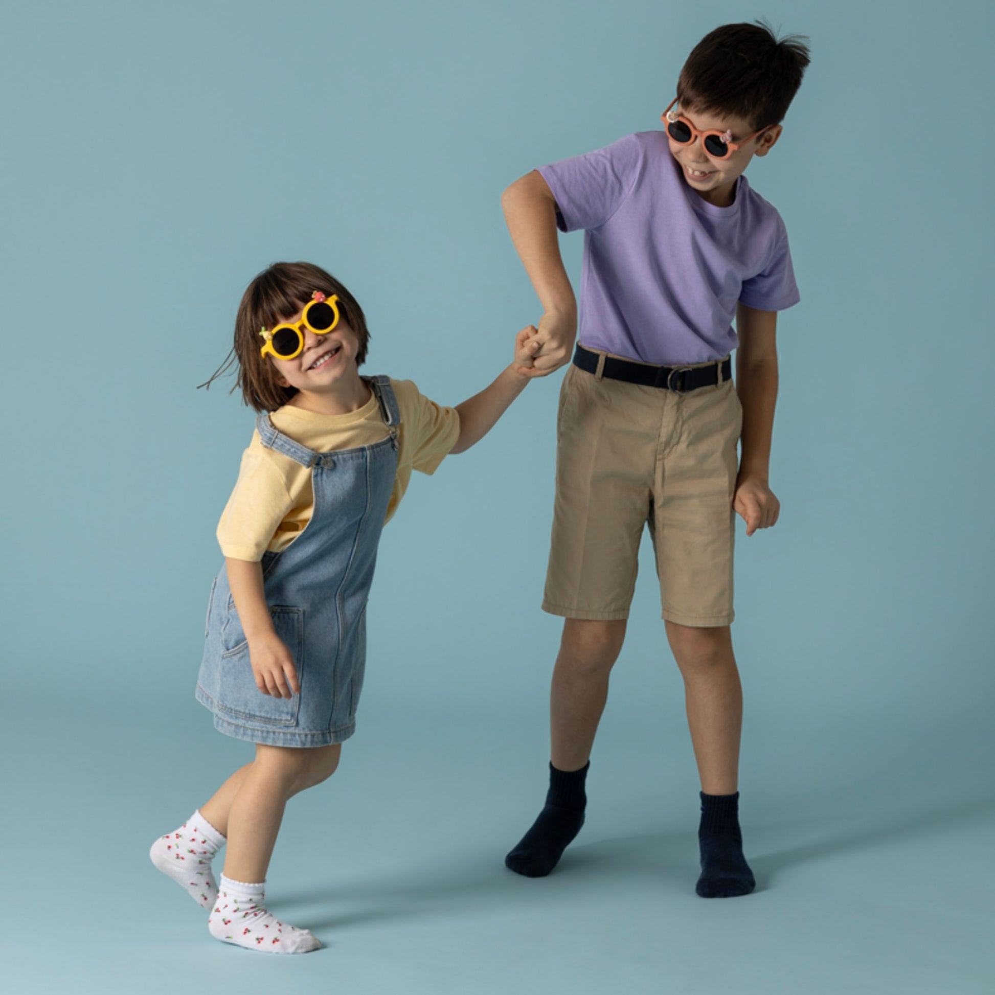 Dwoje dzieci bawiących się w okulary przeciwsłoneczne dla dzieci, ubrane casualowo na niebieskim tle