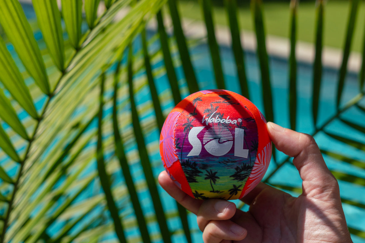Kolorowa piłeczka Waboba Sol trzymana w dłoni na tle zielonych liści i basenu