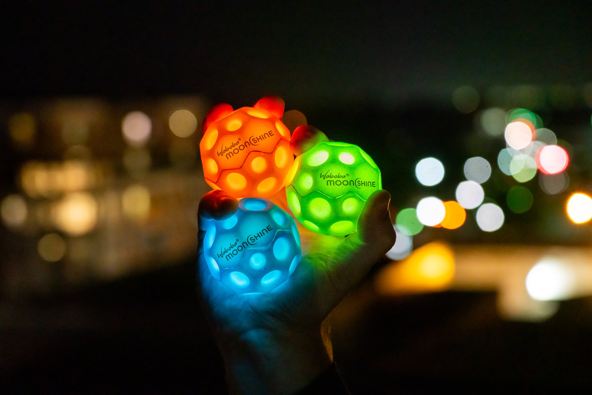Trzy świecące piłeczki Waboba Moonshine w kolorach czerwonym, zielonym i niebieskim trzymane w dłoni na tle nocnego miasta