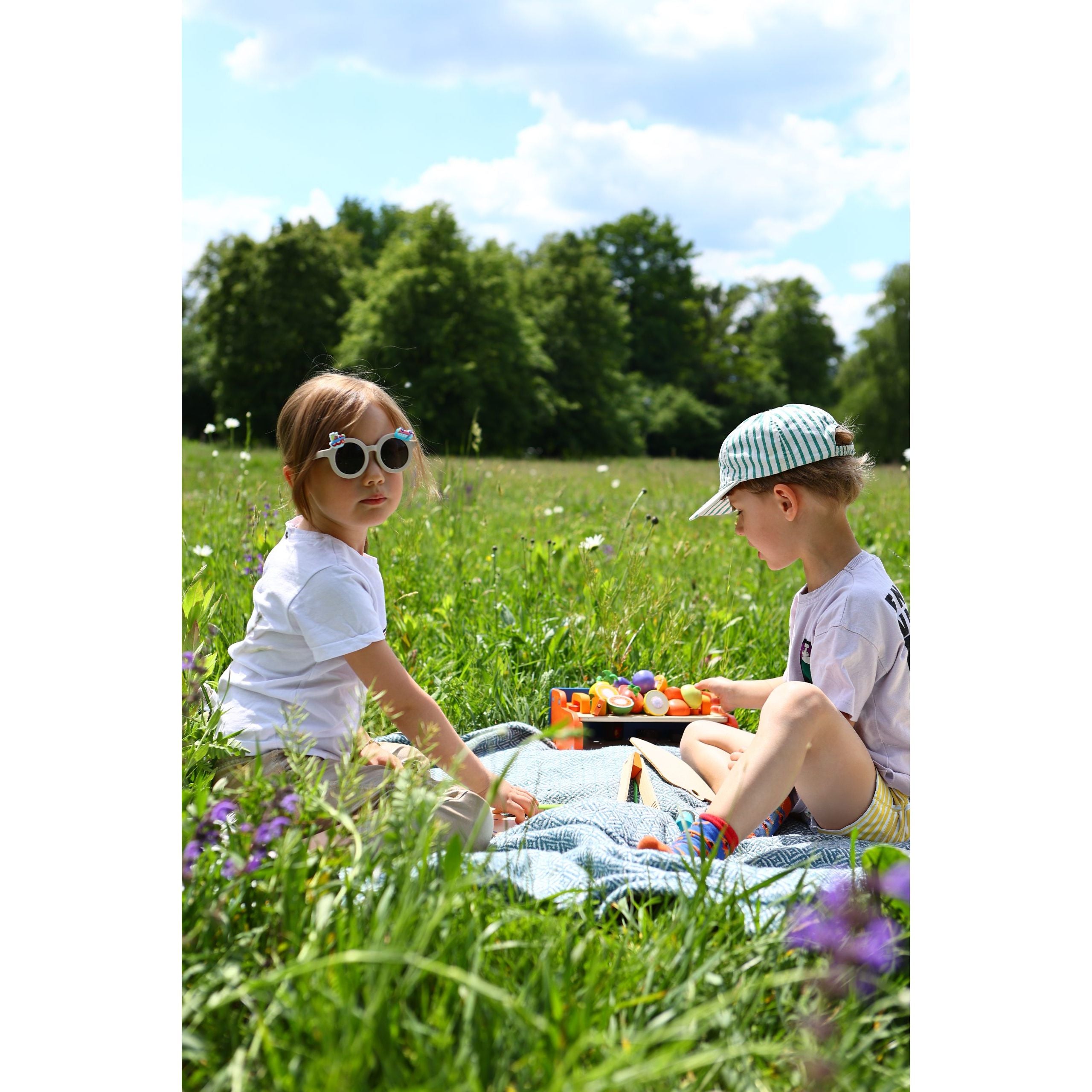 Dwoje dzieci bawiących się na łące z drewnianym grillem dla dzieci na kocu