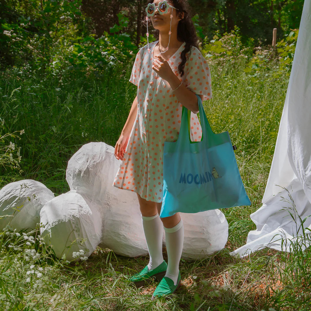 Kobieta w sukience trzymająca niebieską torbę LOQI Moomin na tle zielonej łąki