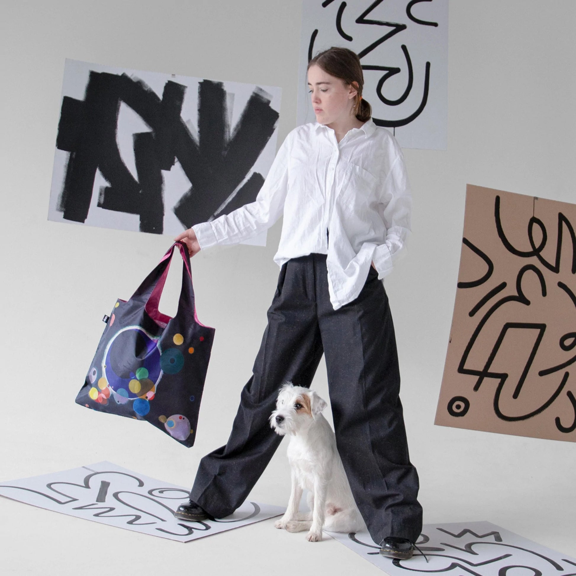 Torba podręczna Wassily Kandinsky w geometryczne wzory trzymana przez kobietę obok psa na tle artystycznych plakatów