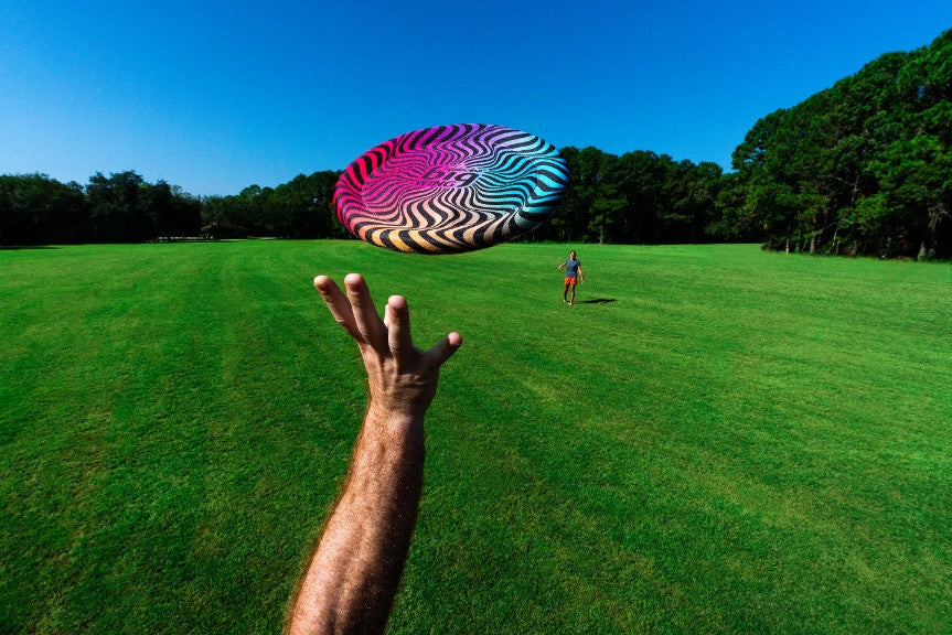 Latający dysk Waboba Bigwing rzucany na zielonym polu z widoczną osobą czekającą na złapanie