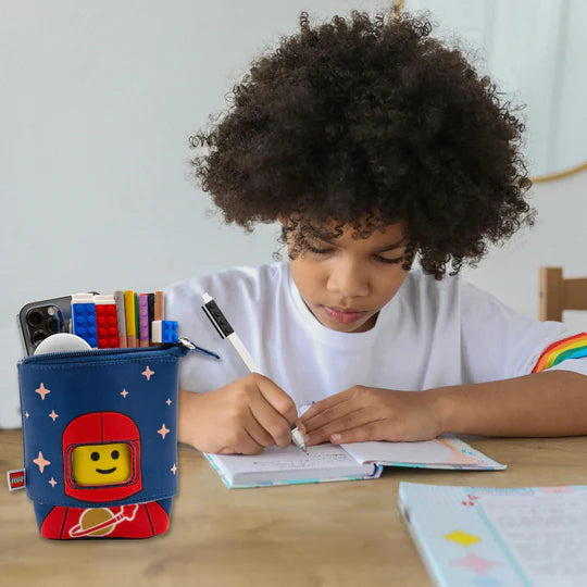 Piórnik LEGO czerwony astronauta z przyborami szkolnymi stojący na biurku obok dziecka piszącego w zeszycie