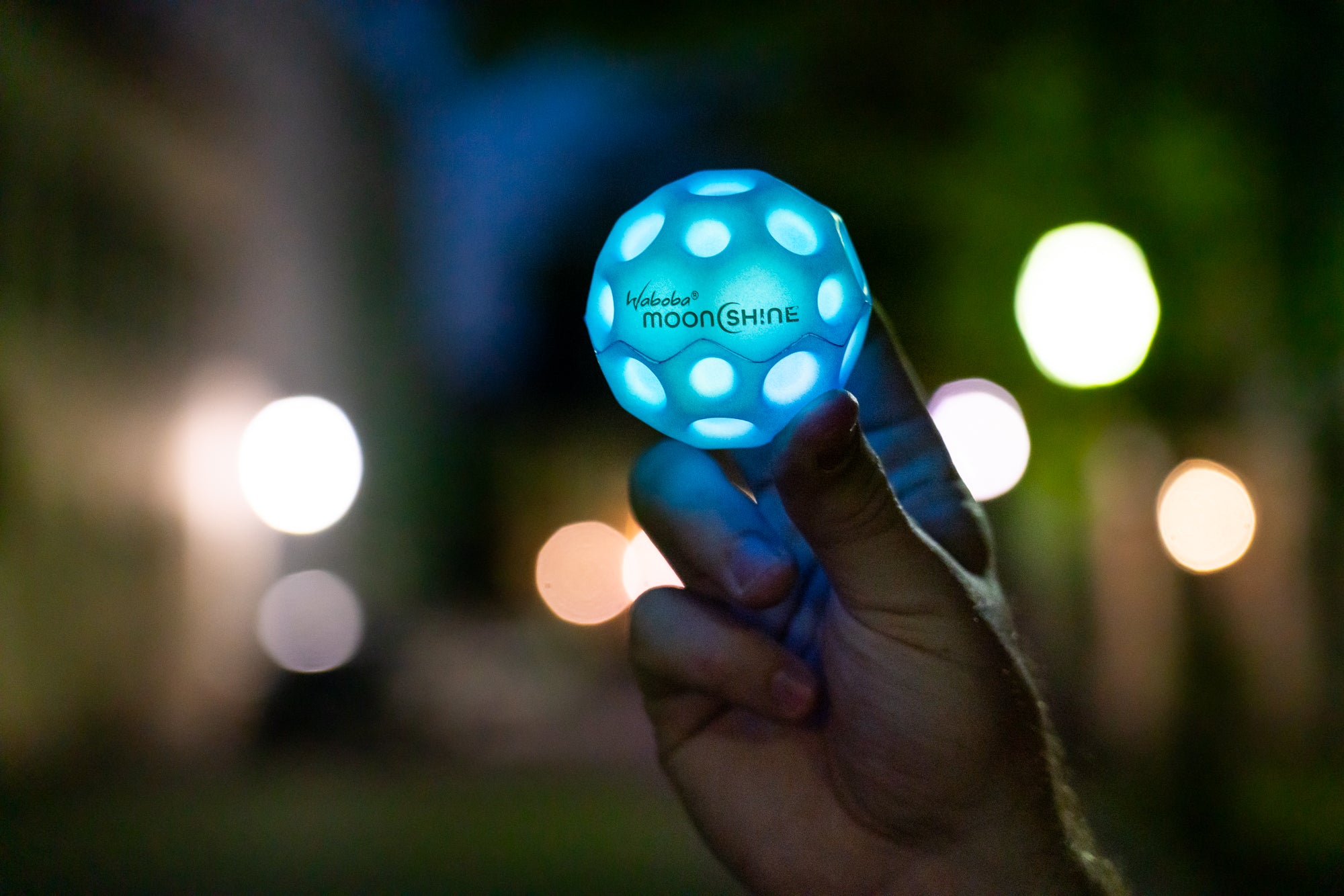 Ręka trzymająca świecącą piłeczkę Waboba Moonshine niebieską na rozmytym nocnym tle