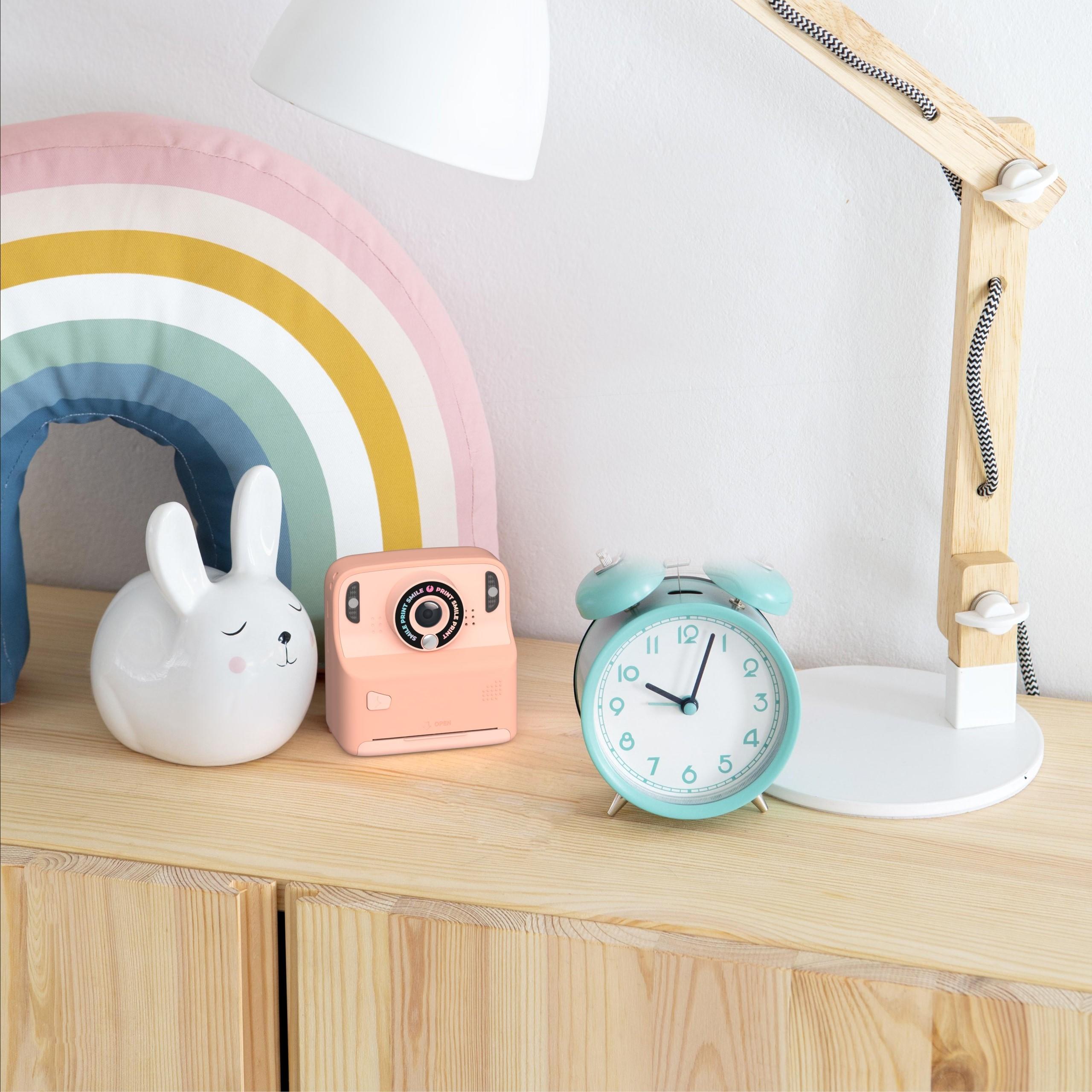 Różowy aparat cyfrowy z drukarką na drewnianej półce obok budzika i dekoracji tęczy