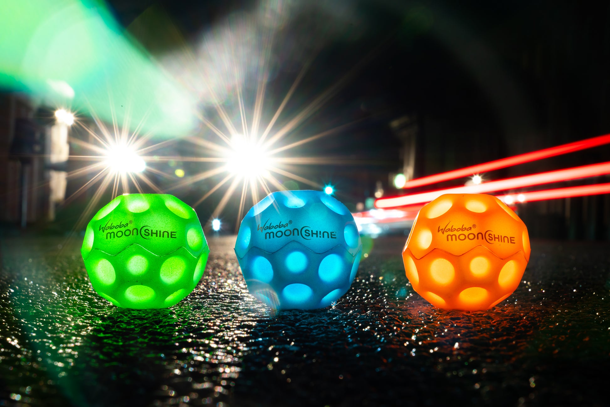 Trzy kolorowe piłeczki świecące Waboba Moonshine w zielonym, niebieskim i pomarańczowym kolorze na mokrej nawierzchni nocą