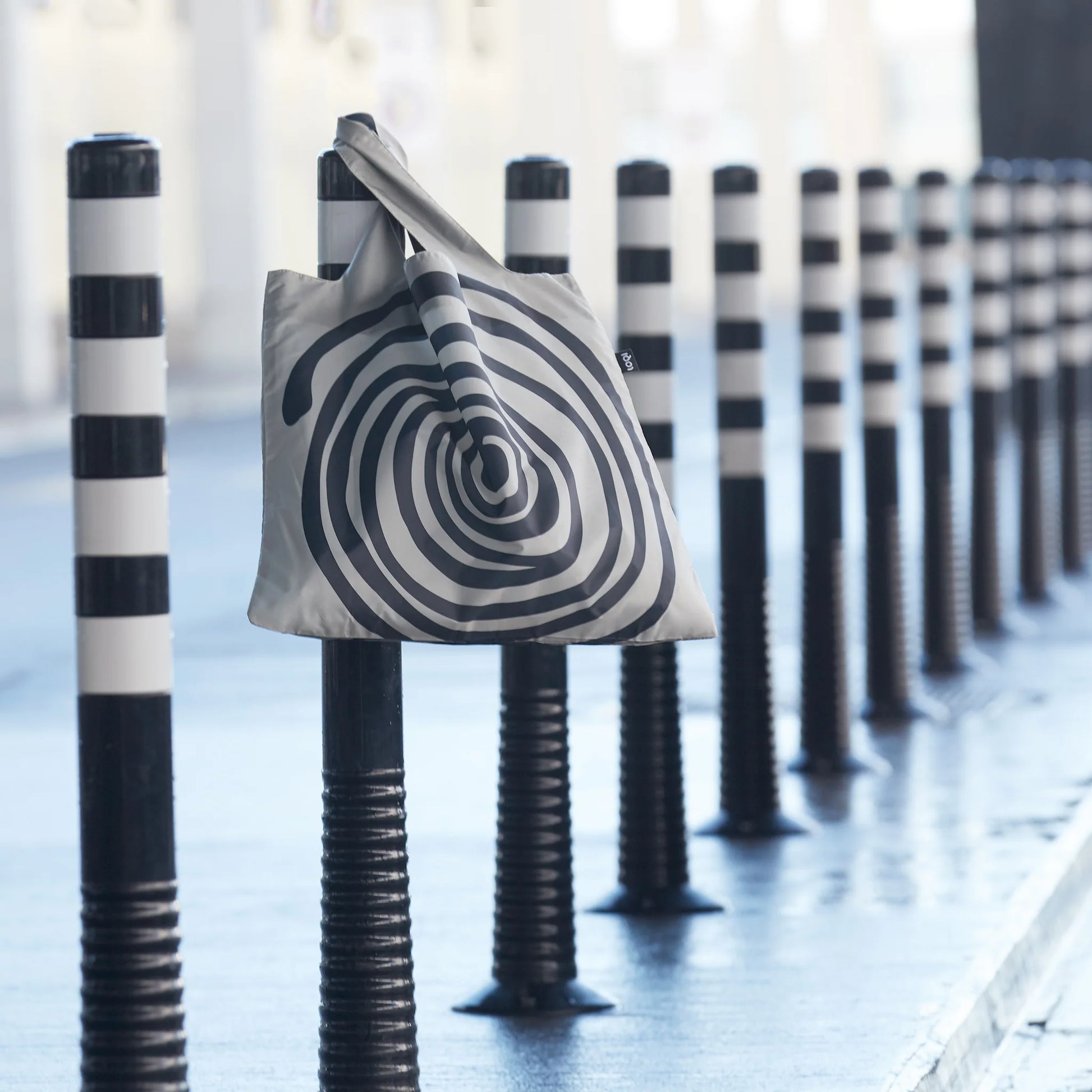 Torba LOQI Louise Bourgeois z czarno-białym wzorem spiralnym zawieszona na czarno-białym słupku na ulicy