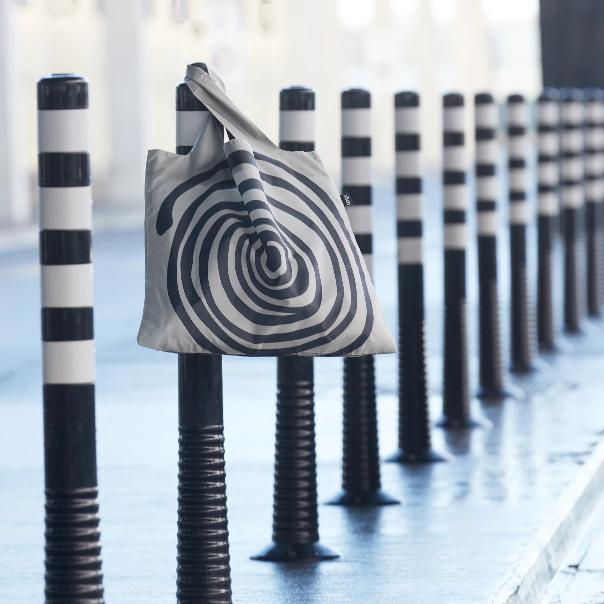 Torba LOQI Louise Bourgeois z czarno-białym wzorem spiralnym zawieszona na czarno-białym słupku na ulicy