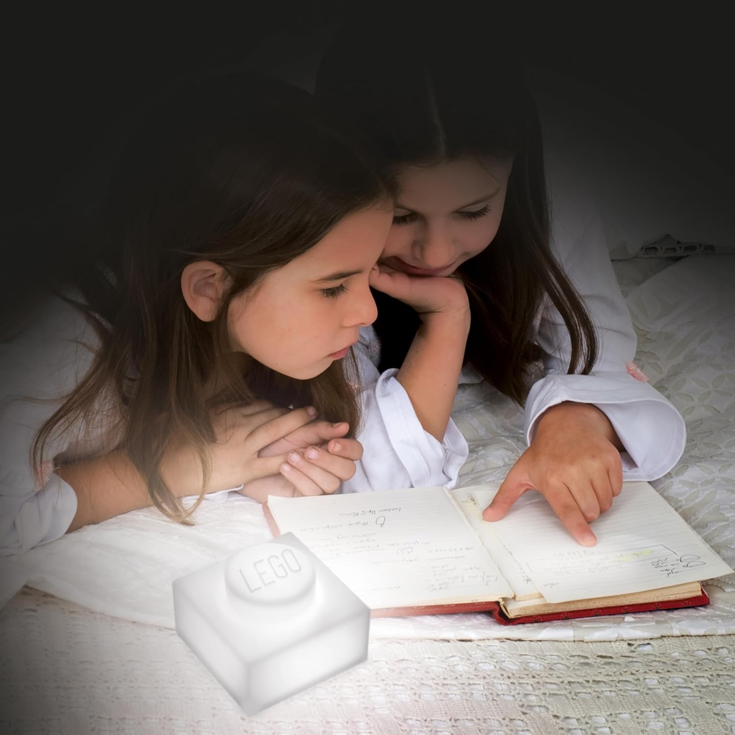 Dwie dziewczynki czytające książkę obok silikonowej lampki LEGO w kształcie klocka