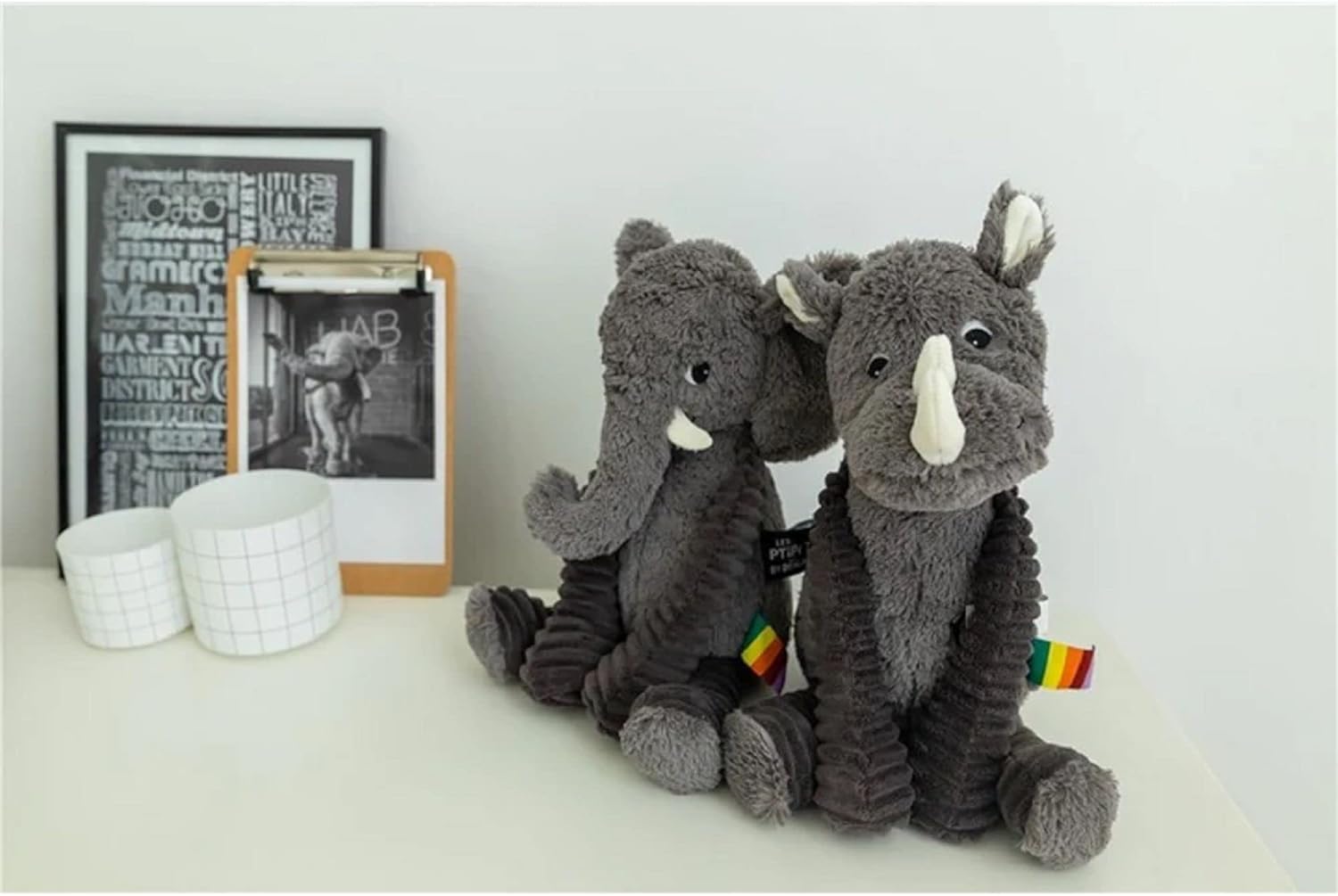Pluszowy słoń szary siedzący na białym stole obok ramki ze zdjęciem i dwoma kubkami