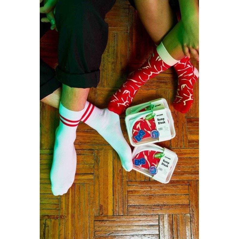 Kolorowe skarpetki steak dla dorosłych w białe i czerwone wzory prezentowane na stopach dwóch osób obok opakowań mięsa EMS
