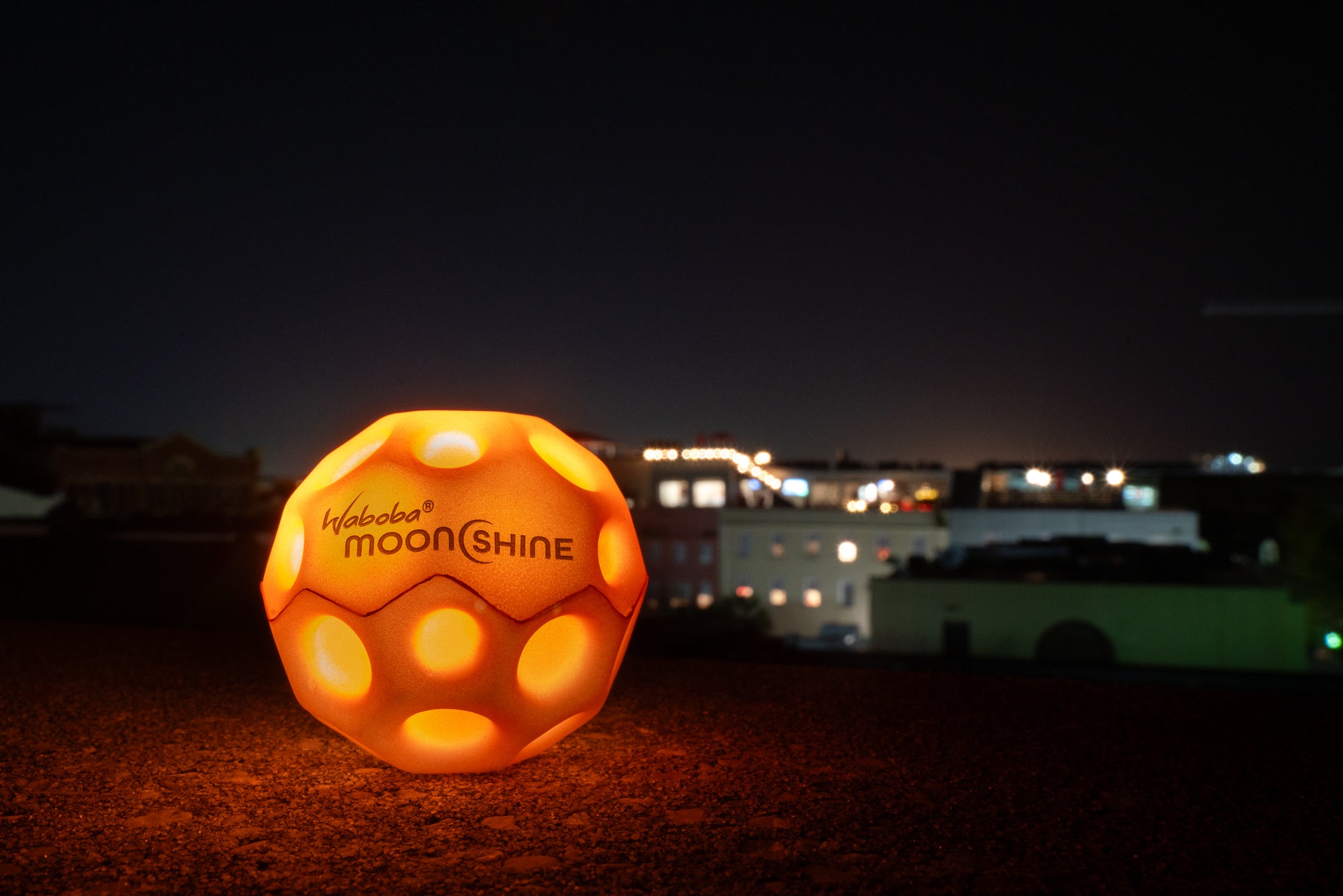 Pomarańczowa piłeczka świecąca Waboba Moonshine świecąca w nocy na tle miasta
