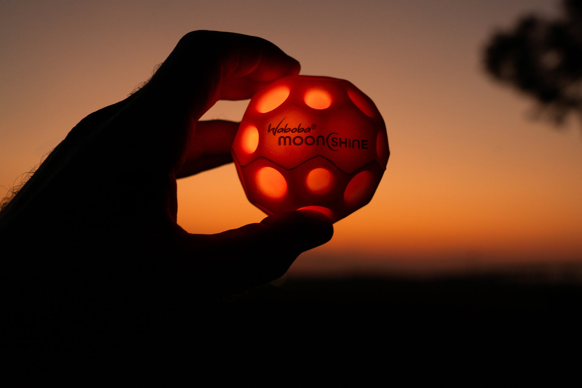 Czerwona piłeczka świecąca Waboba Moonshine trzymana w dłoni na tle zachodu słońca
