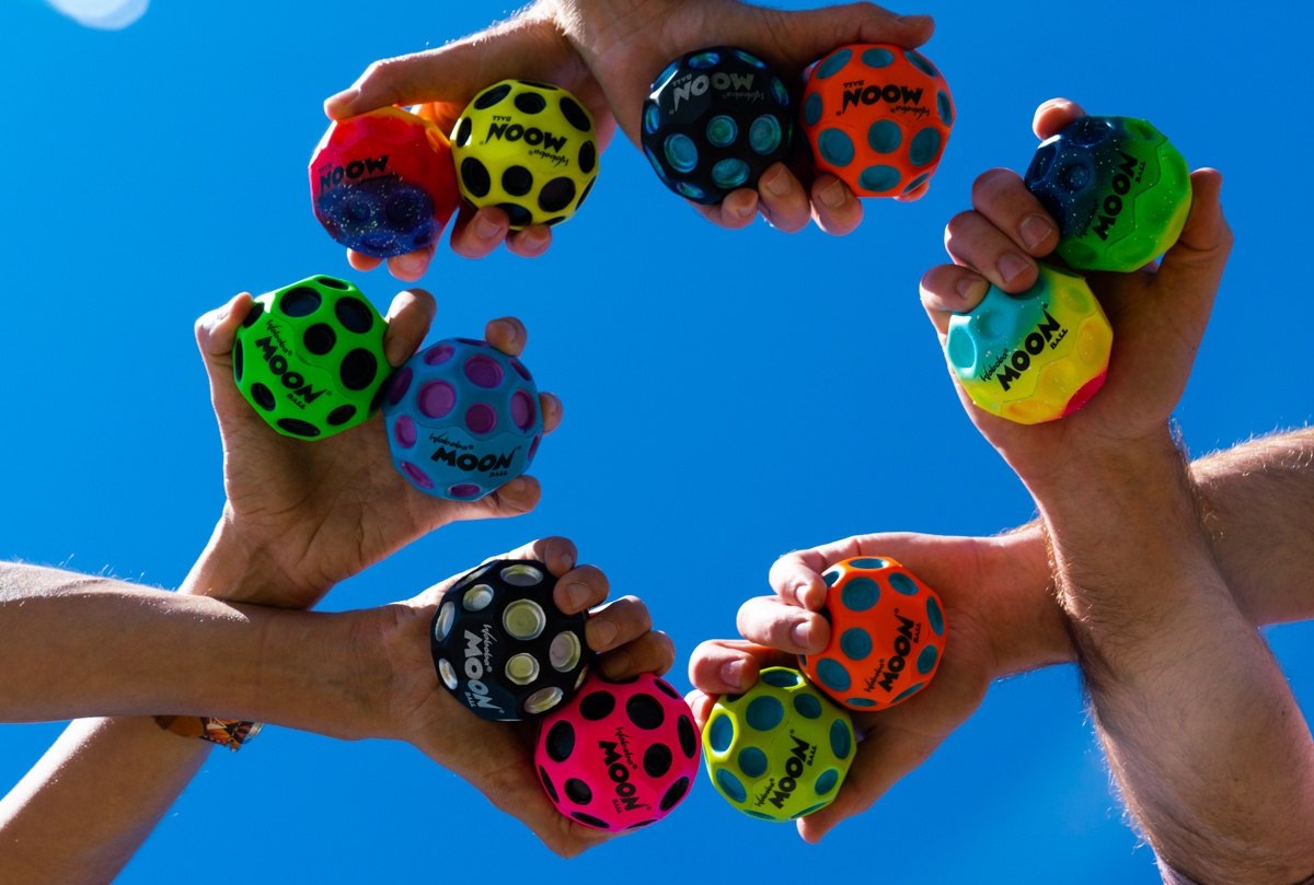 Kolorowe piłeczki Waboba Moon Ball trzymane w dłoniach na tle niebieskiego nieba z piłeczką Waboba Moon Ball niebieską