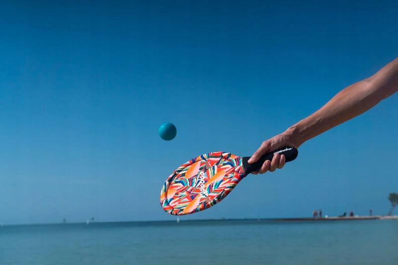 Paletki plażowe Waboba z kolorowym wzorem trzymane przez osobę na tle morza i niebieskiego nieba