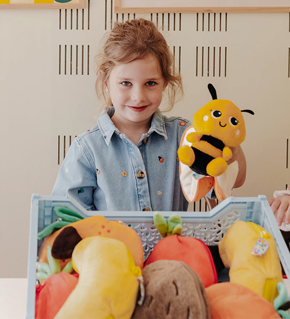 Dziewczynka trzymająca pluszaka pszczoła mandarynka w jasnej koszuli z zabawkami owocowymi w skrzyni