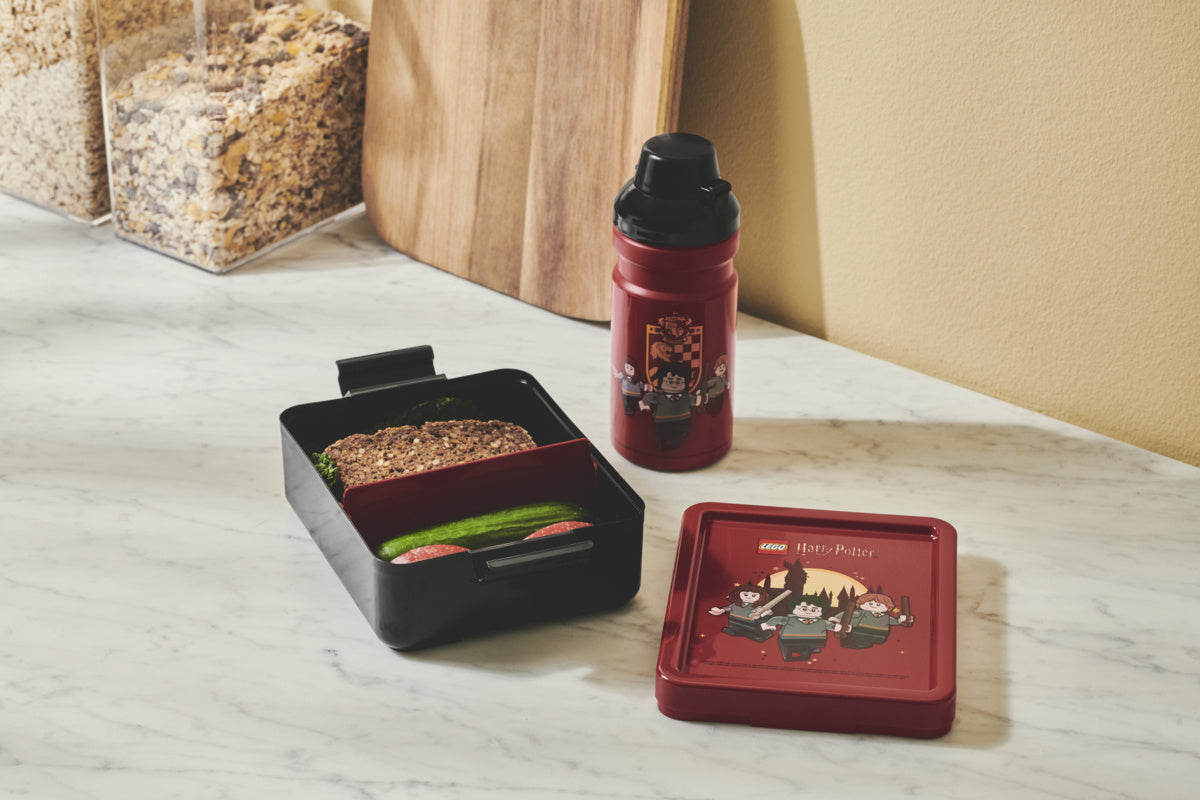 Bidon LEGO Harry Potter Gryffindor w czerwonym kolorze z motywem czarodziejów obok lunchboxa z kanapką i warzywami