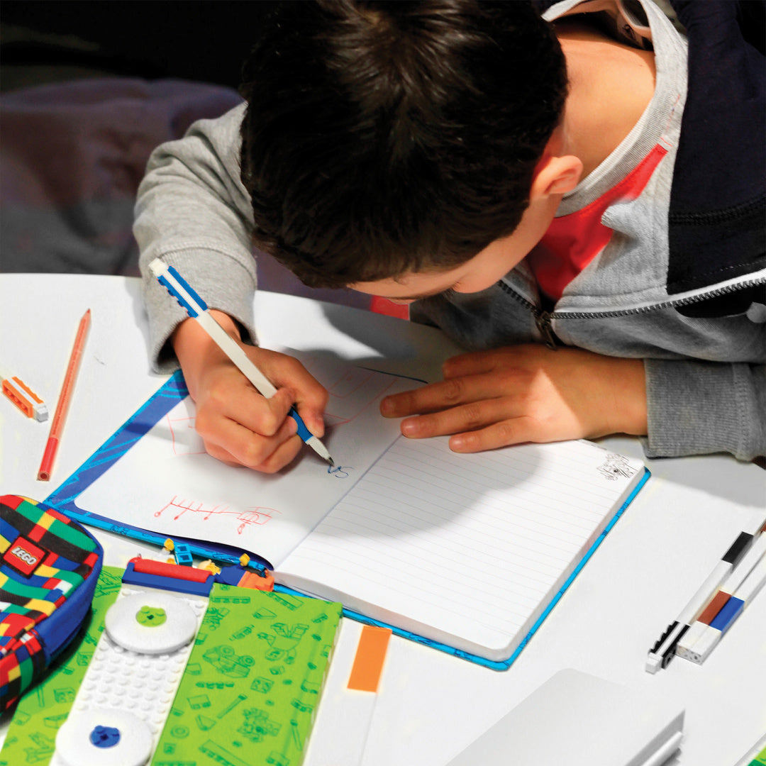 Chłopiec piszący w notesie długopisem żelowym LEGO na białym biurku z kolorowymi przyborami szkolnymi