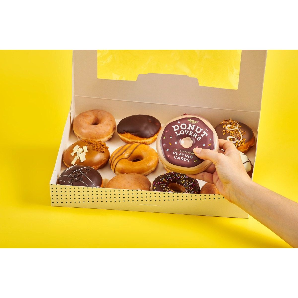 Karty do gry z motywem donutów w pudełku obok różnorodnych lukrowanych pączków na żółtym tle