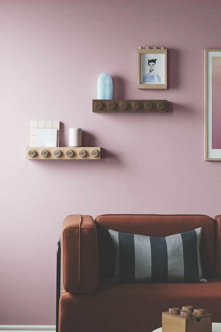 Drewniana półka LEGO w naturalnym i ciemnym kolorze zamontowana na różowej ścianie nad czerwoną kanapą