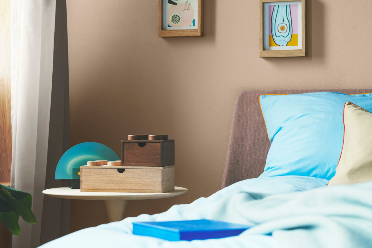 Drewniana szufladka LEGO na biurko w naturalnym i ciemnym kolorze ustawiona na stoliku obok łóżka