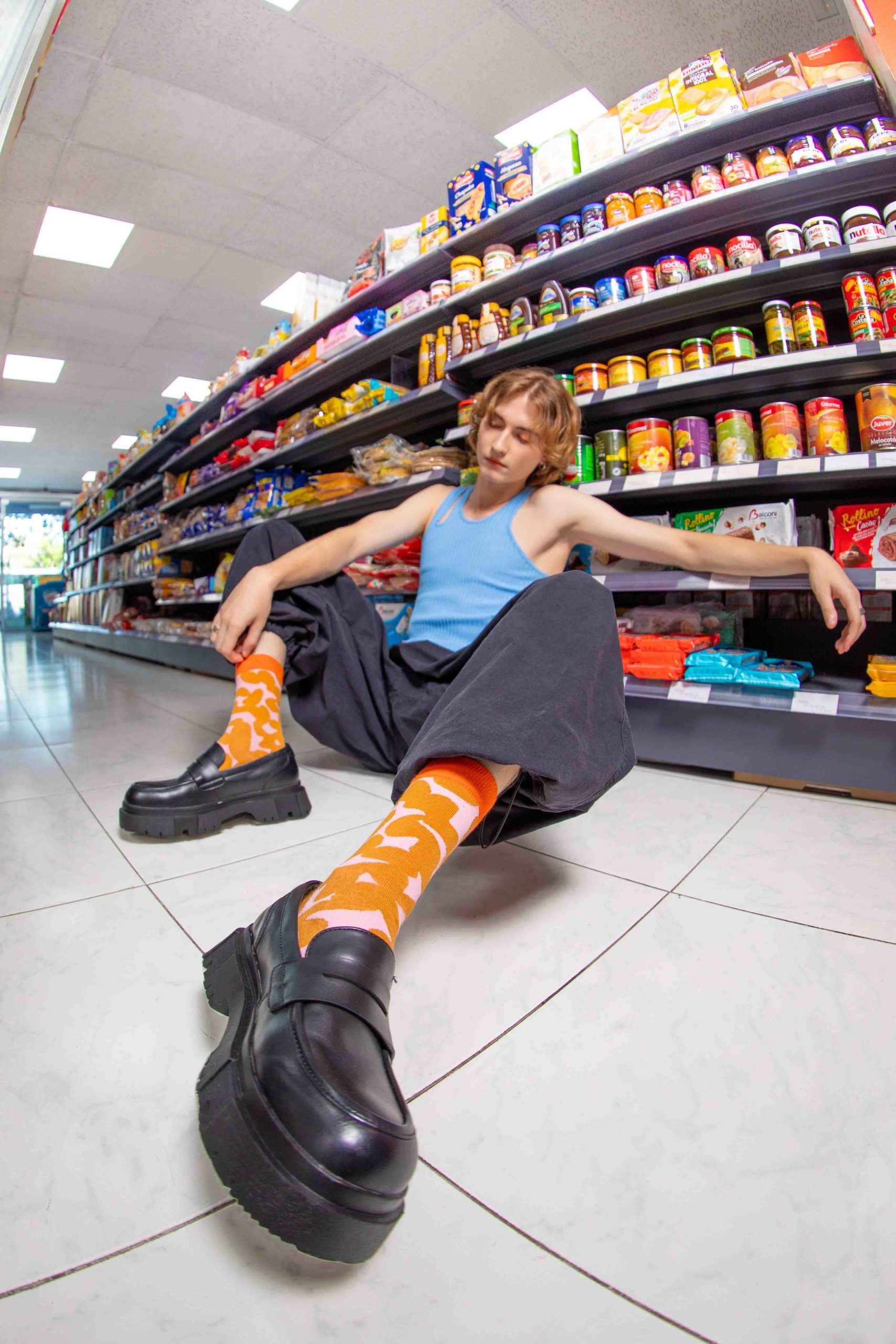 Młoda osoba w pomarańczowych skarpetkach peanut butter siedzi na podłodze sklepu spożywczego obok półek z produktami
