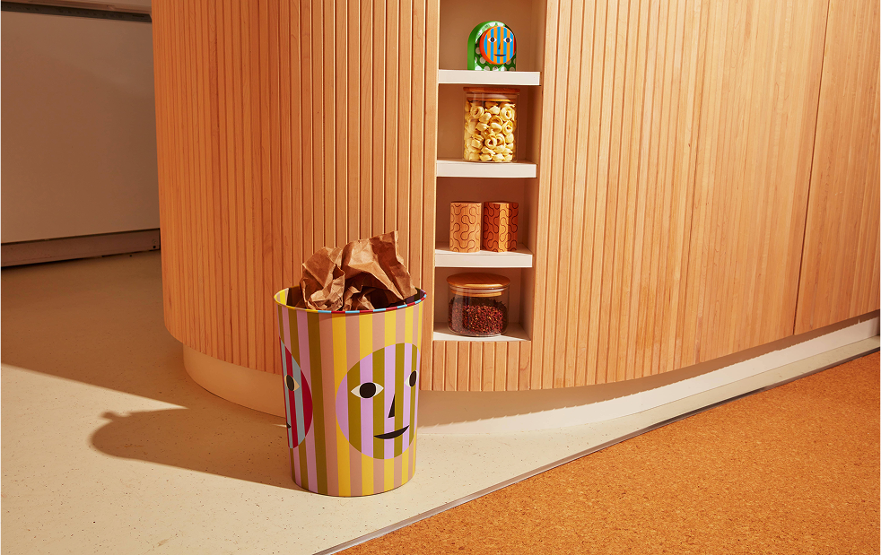 Kolorowy kosz na śmieci design Areaware z motywem twarzy ustawiony przy drewnianej ścianie