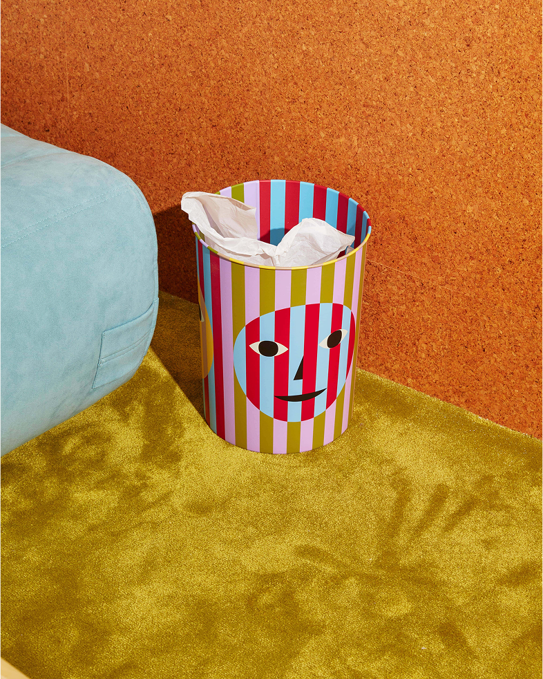 Kolorowy kosz na śmieci design Areaware z wzorem twarzy w paski obok niebieskiej sofy