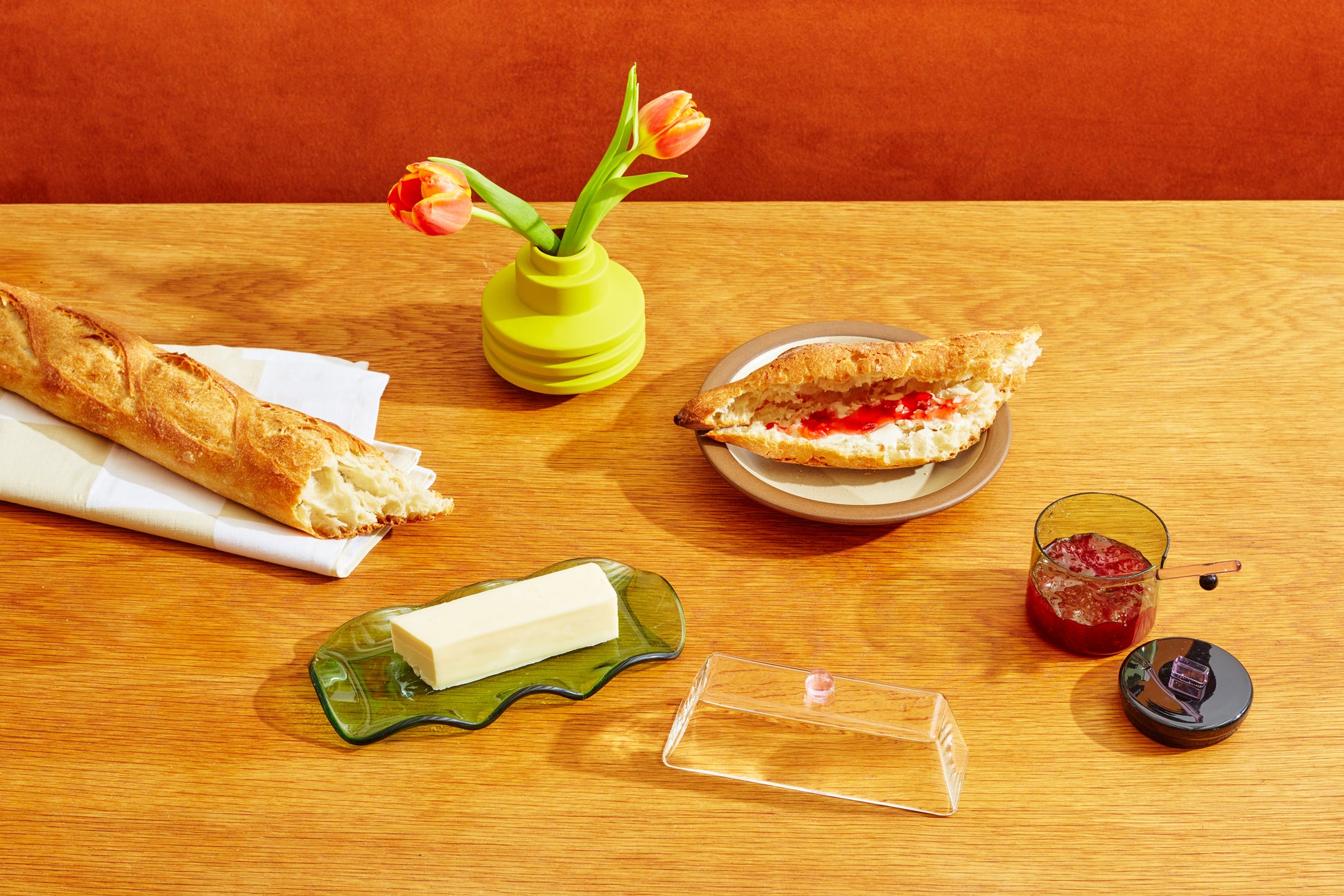 maselniczka szklana zielono-różowa z masłem na drewnianym stole obok bagietki i dżemu