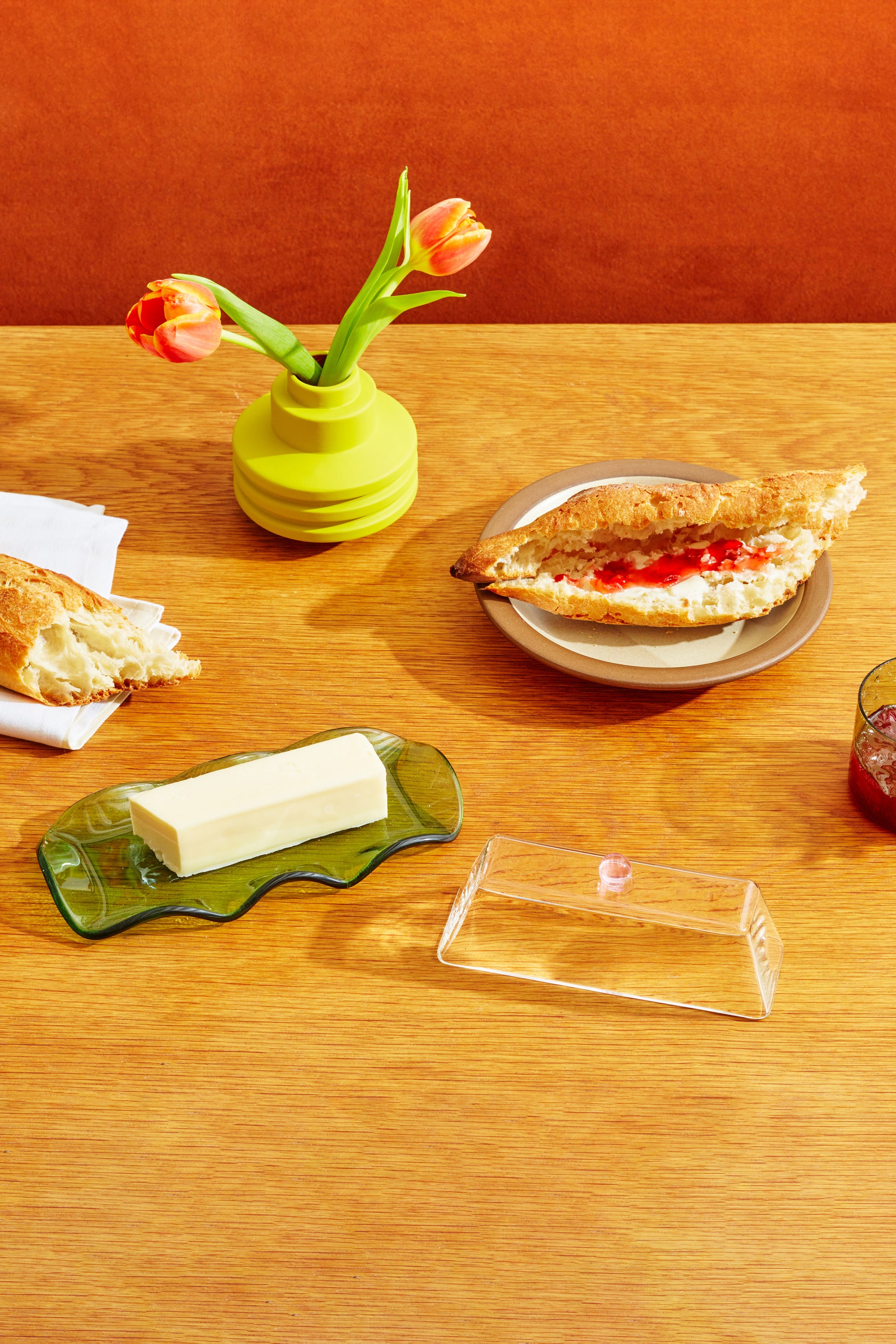 Maselniczka szklana zielono-różowa z kostką masła na drewnianym stole obok pieczywa i wazonu z tulipanami