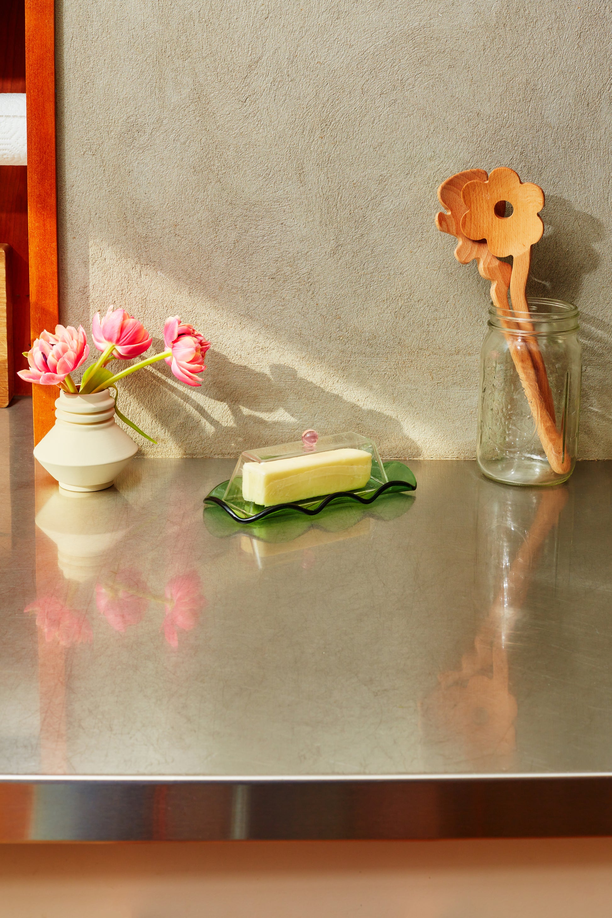 Maselniczka szklana zielono-różowa z masłem na metalowym blacie obok kwiatów i drewnianych łyżek