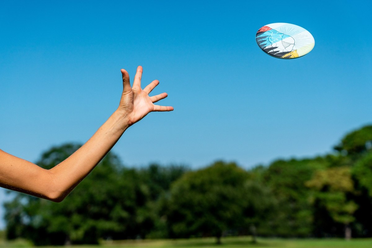 Ręka rzucająca frisbee Waboba Wingman na tle nieba i zielonych drzew w parku