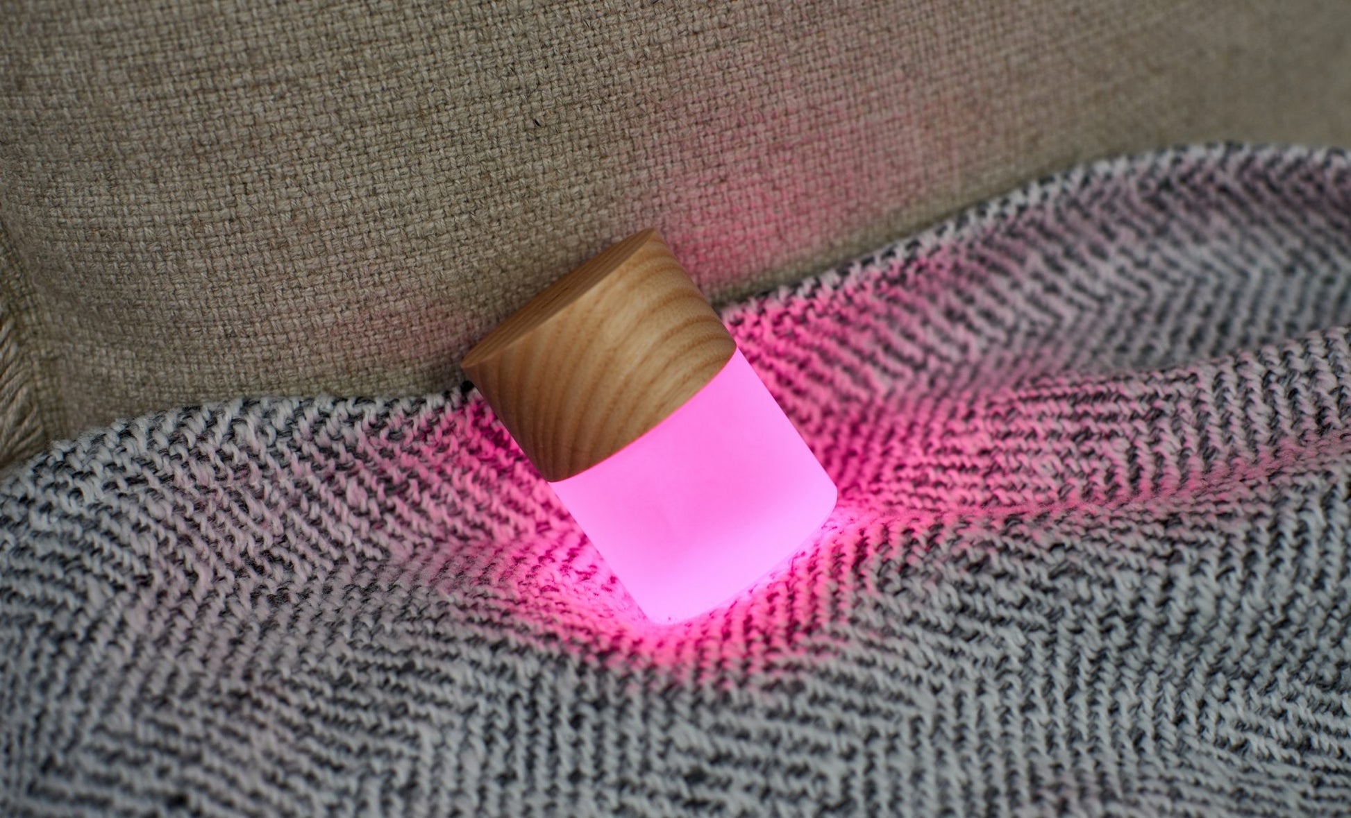 Lampka nocna z drewna jesionowego świecąca na różowo na tle szarego materiału i beżowej kanapy