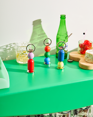 Kolorowy otwieracz do butelek design Areaware ustawiony na zielonym blacie obok napojów i przekąsek