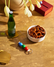 Otwieracz do butelek Abaco Areaware z kolorowymi kulkami na stole obok butelki, miski z precelkami i kwiatów