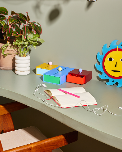 Kolorowy pojemnik Cache Box Areaware na biurku obok notatnika, rośliny i zegara słonecznego