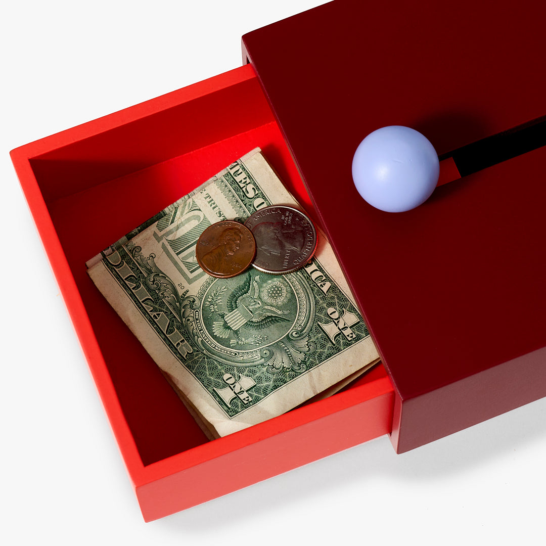 Pojemnik Cache Box Areaware w kolorze czerwonym z granatową pokrywką i kulkowym uchwytem, z pieniędzmi w środku