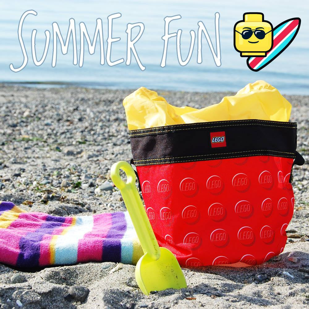 Kubełek LEGO czerwony z czarnym rantem na plaży z żółtą łopatką i kolorowym ręcznikiem