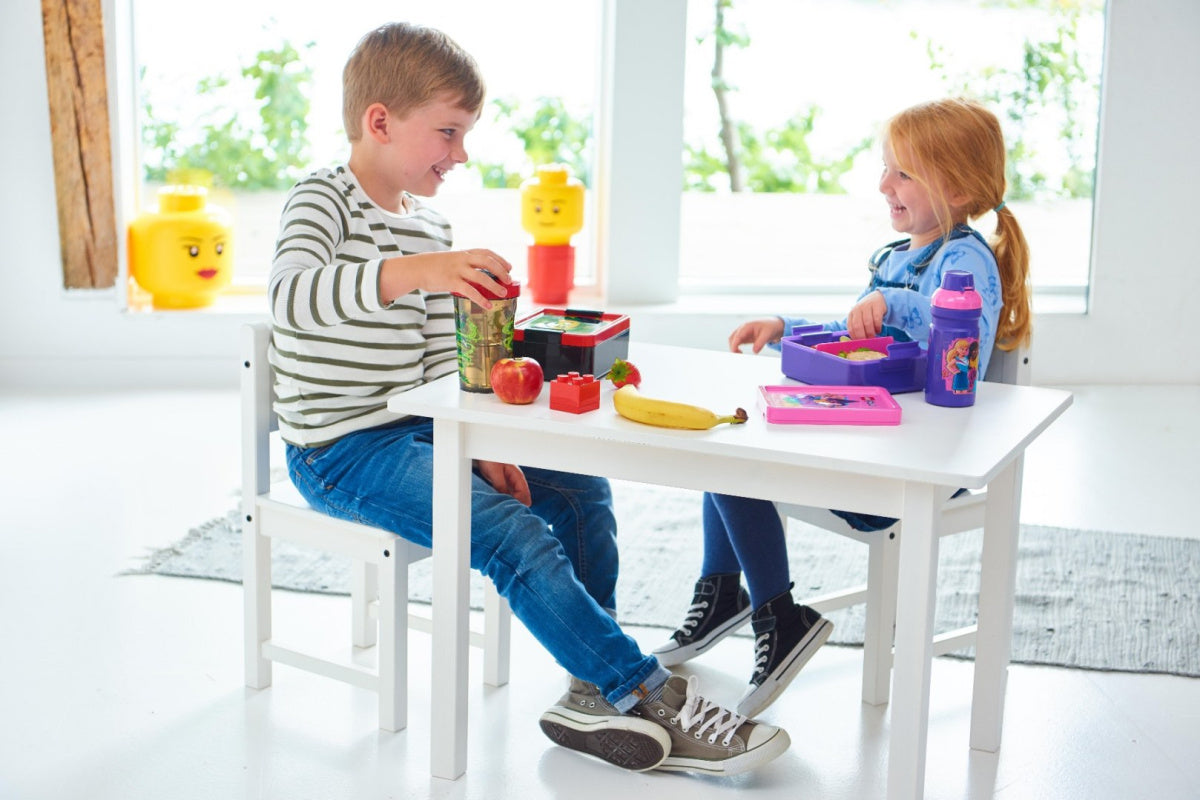 Dwoje dzieci jedzących przy białym stoliku z lunchbox LEGO Friends i owocami w jasnym pokoju