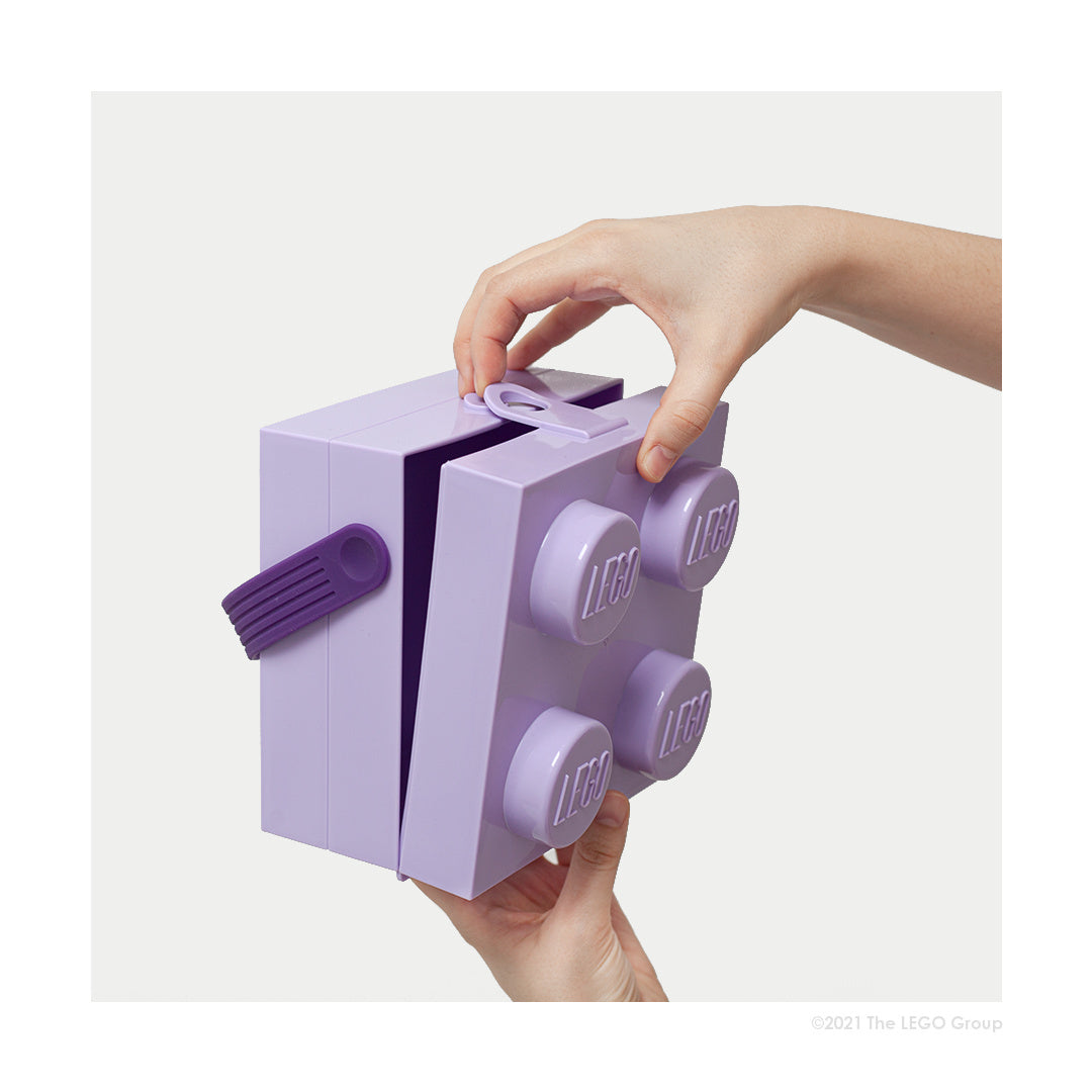 Lunchbox LEGO fioletowy w kształcie klocka trzymany w dłoniach z otwieranym wieczkiem