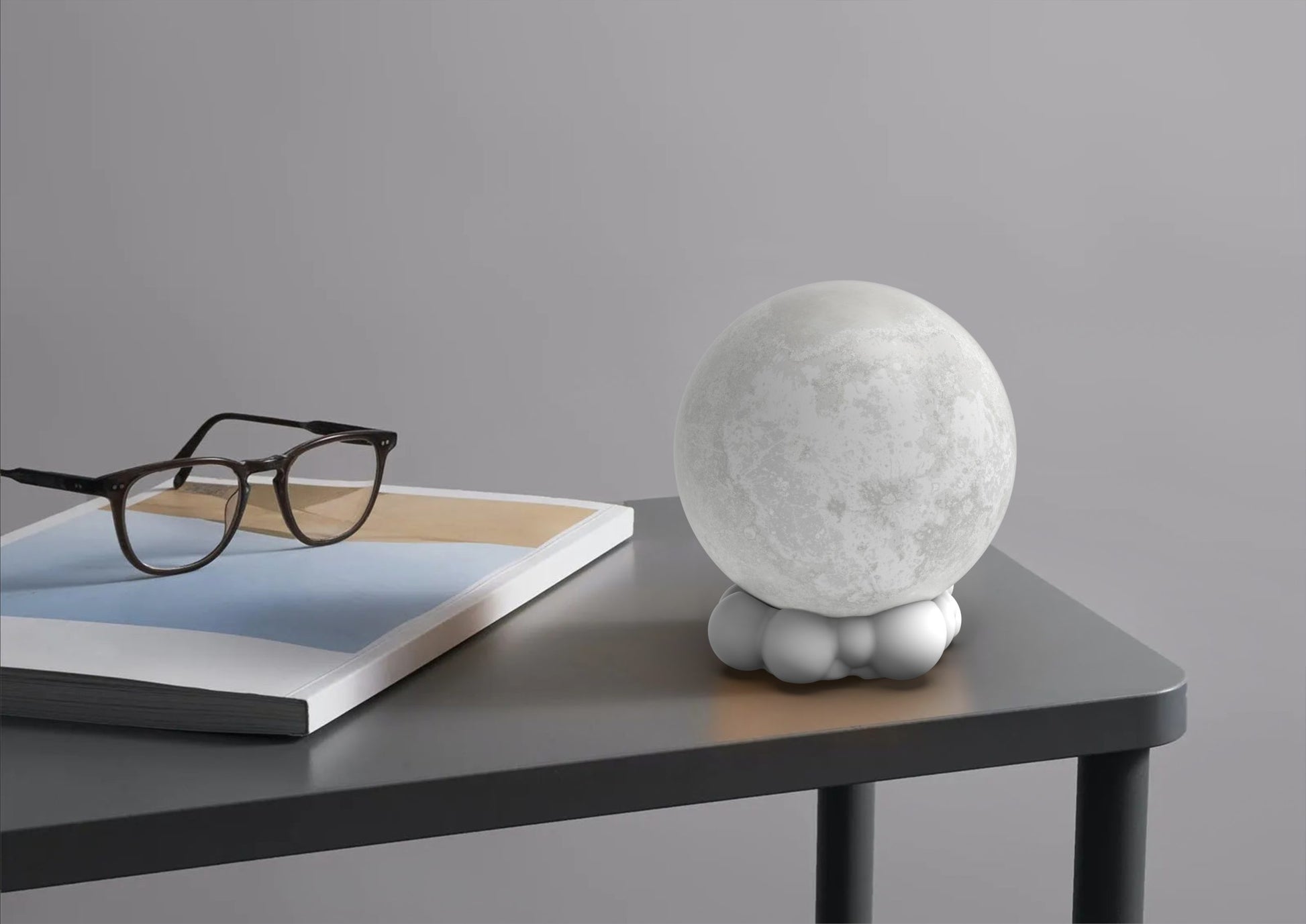 Lampka nocna z głośnikiem Yamann w kształcie księżyca na szarym stole obok okularów i książki