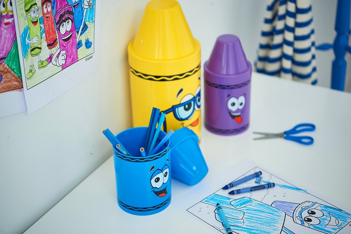 Fioletowy pojemnik na kredki Crayola fioletowy obok niebieskiego i żółtego pojemnika na biurku dziecięcym