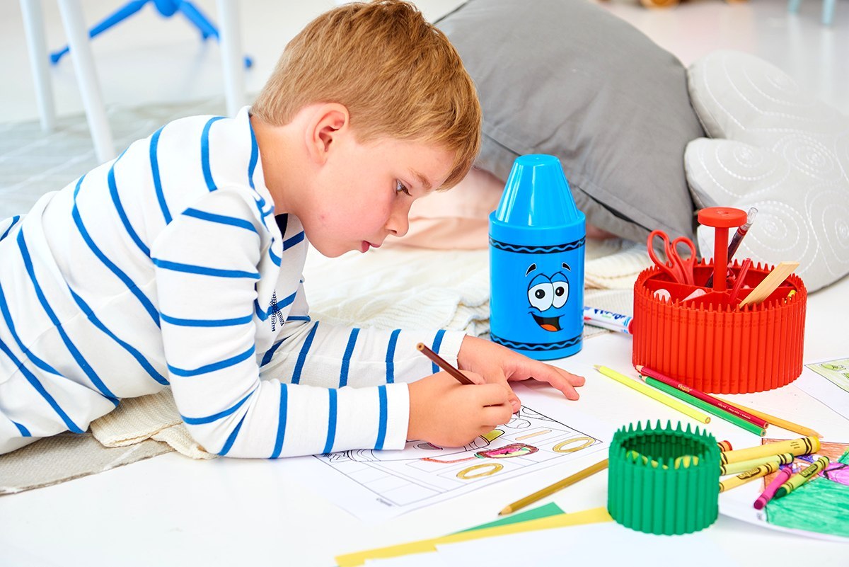 Chłopiec rysujący kredkami obok fioletowego pojemnika na kredki Crayola fioletowy i innych akcesoriów plastycznych