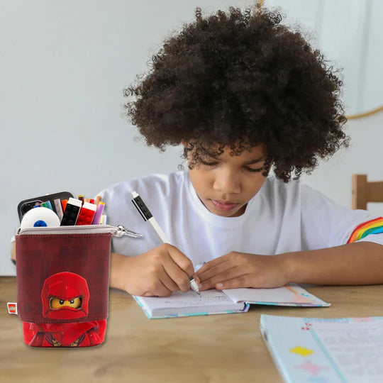 piórnik LEGO Ninjago Kai w czerwonym kolorze z nadrukiem postaci Kai na biurku obok dziecka piszącego w zeszycie
