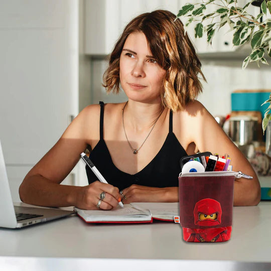 Piórnik LEGO Ninjago Kai w kształcie kubka z akcesoriami na biurku obok młodej kobiety piszącej notatki