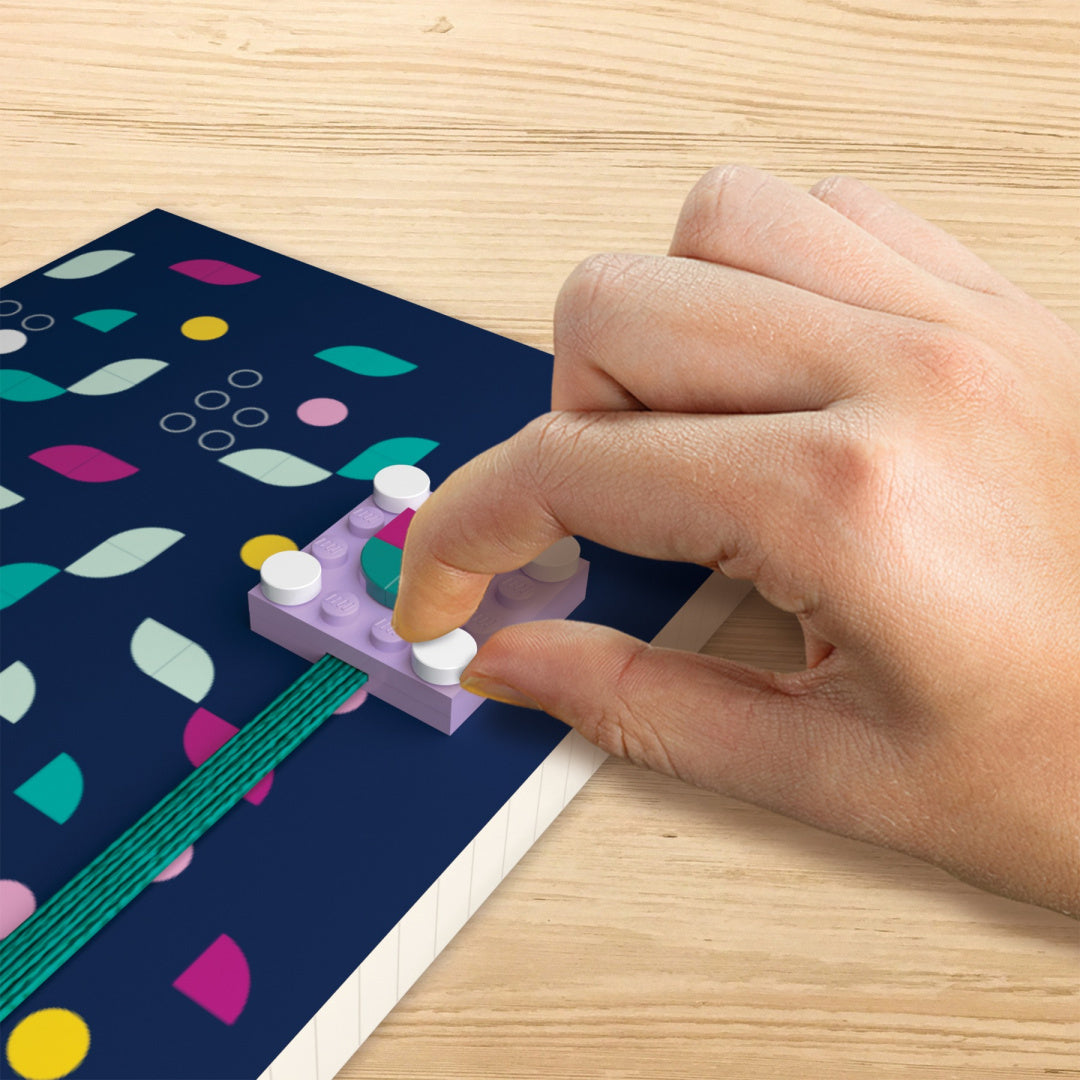 Notatnik LEGO DOTS z kolorową okładką z wzorem liści i klockiem dociskanym przez dłoń na drewnianym stole