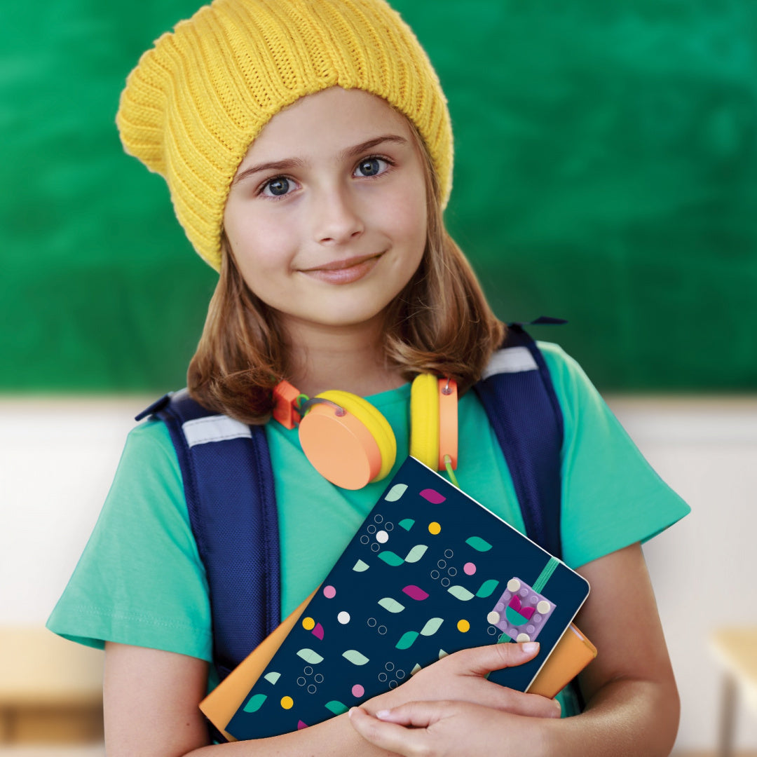 Dziewczynka trzymająca kolorowy notatnik LEGO DOTS w klasie z żółtą czapką i słuchawkami